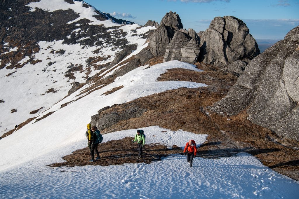 Four hikers climb a slope of snow and brown tundra below jagged granite outcrops, low sun lighting the rock.
