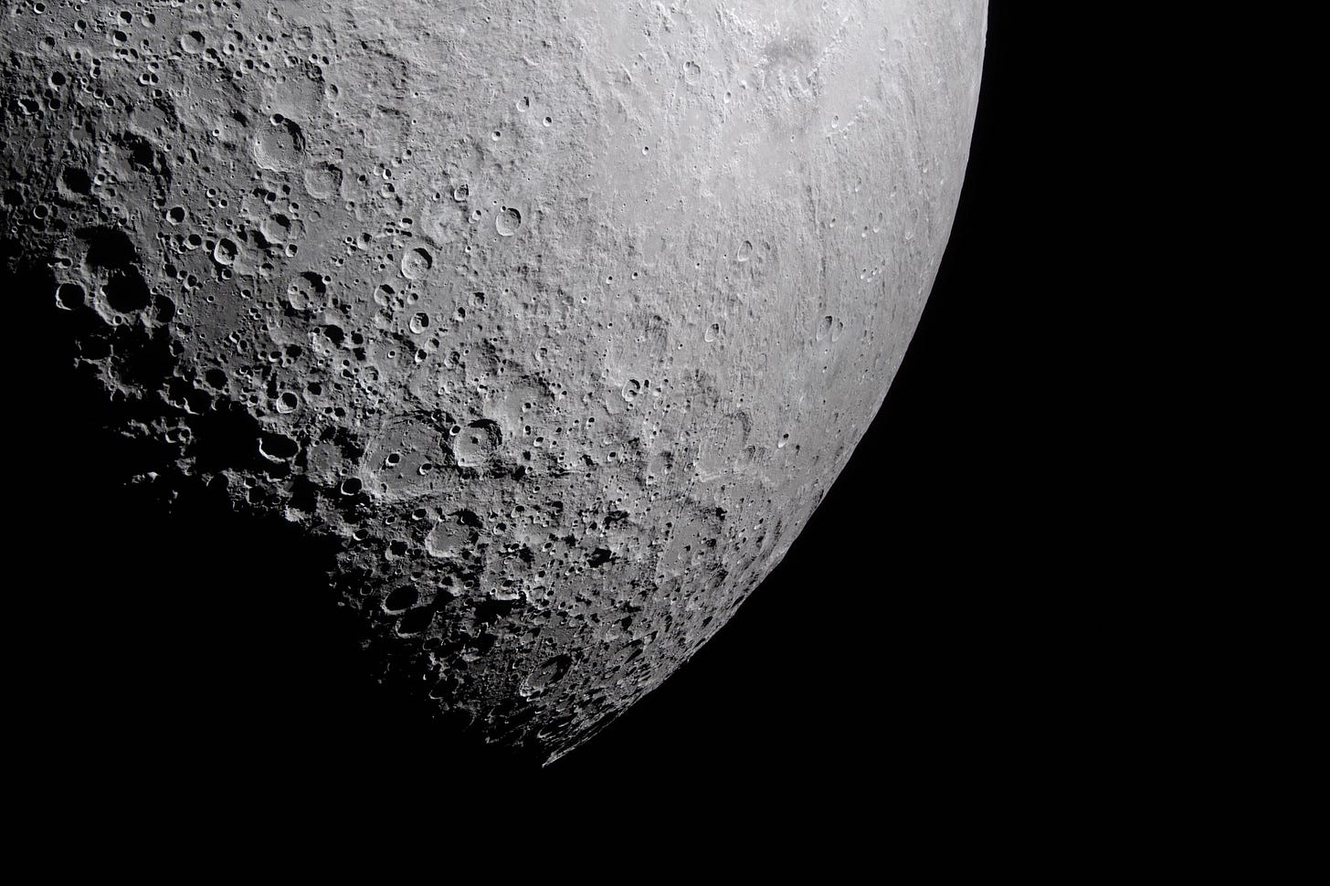 Part of the Moon stands out against the blackness of space. Its surface is marked with many craters of different sizes. The craters are mostly on the left and bottom. The upper right portion of the Moon's surface is much smoother.