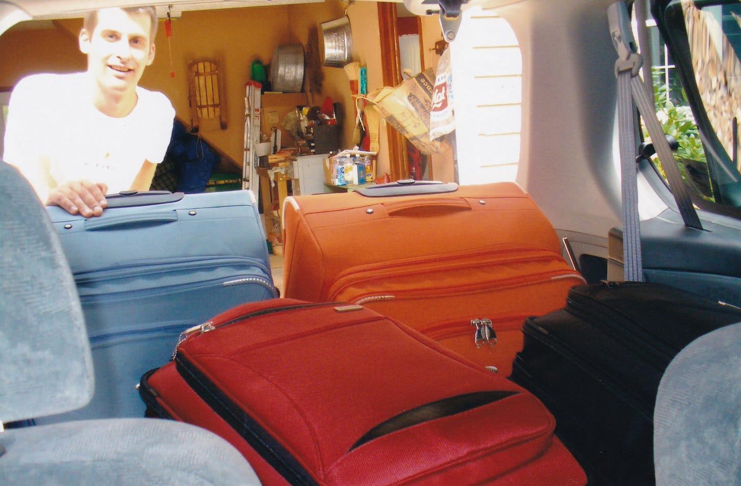 A man looks at the camera. Before him are four big suitcases in the back of a car. It is daytime.