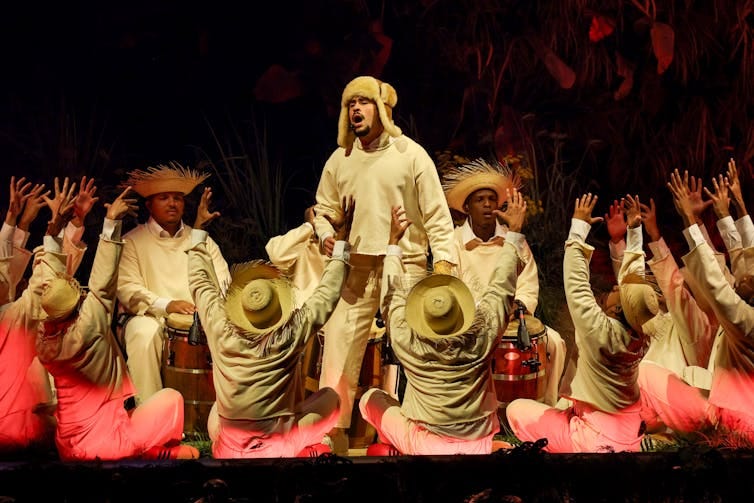 Man in beige clothes and hunting cap sings while surrounded by circle of men wearing straw hats and some playing drums