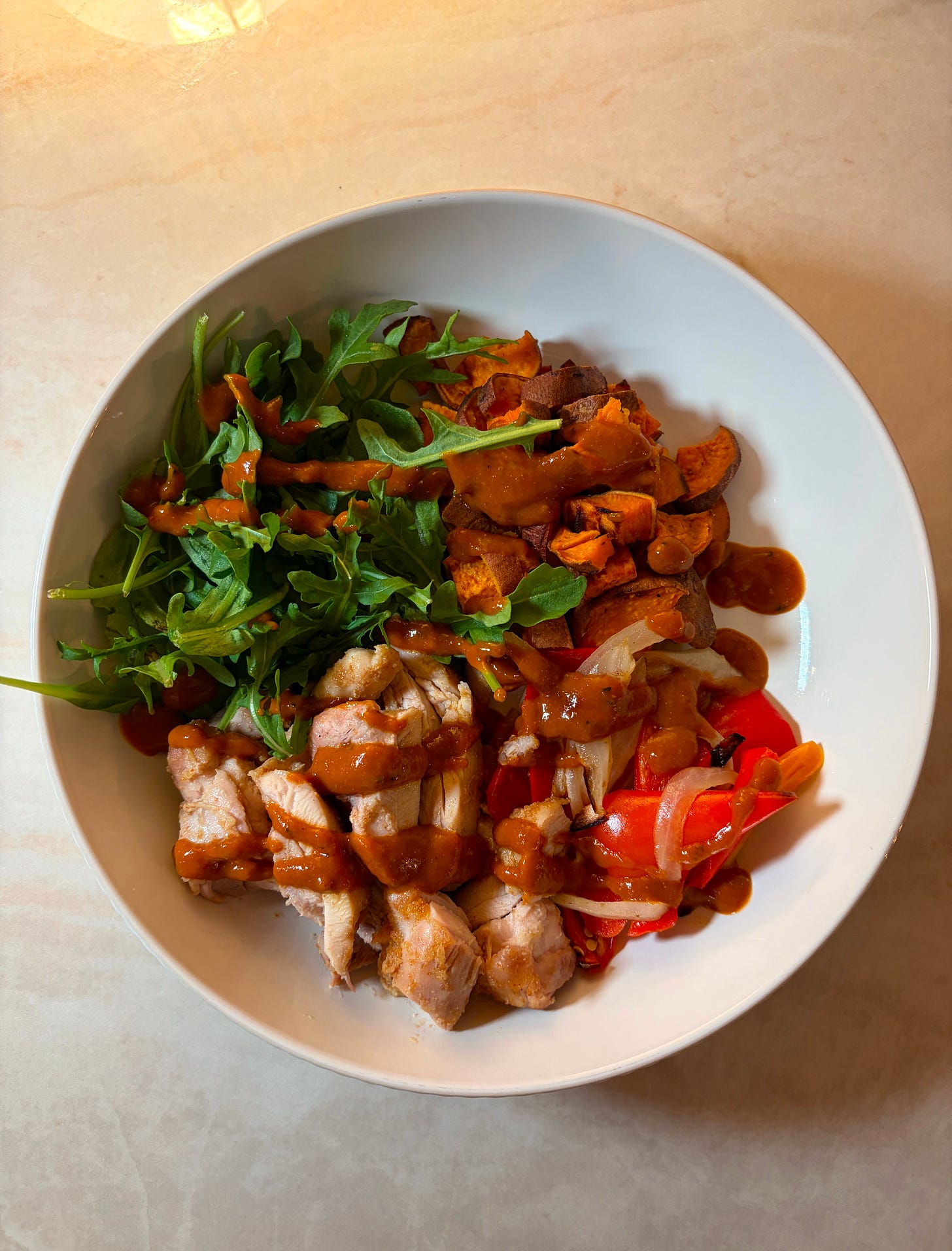 A bowl with a base of arugula topped with sliced BBQ chicken thighs, roasted sweet potato cubes, and caramelized peppers and onions, drizzled with golden BBQ sauce