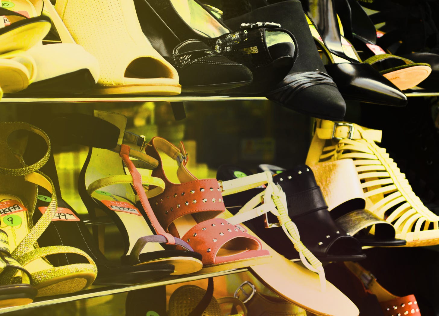 A shelf stacked with thrifted shoes, focusing in one one white and red heel hanging off the edge.