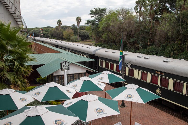 Four photographs from South Africa. Visitors stand on the hand-drawn floor map of old District Six inside the District Six Museum in Cape Town. A steep street of brightly painted houses in Bo-Kaap, with two people walking uphill carrying shopping. A Rovos Rail train alongside a platform at Capital Park station in Pretoria, with green and white branded umbrellas in the foreground. The interior of Clarke's Bookshop in Cape Town, seen from an upstairs landing, with crowded shelves and customers browsing tables of books.