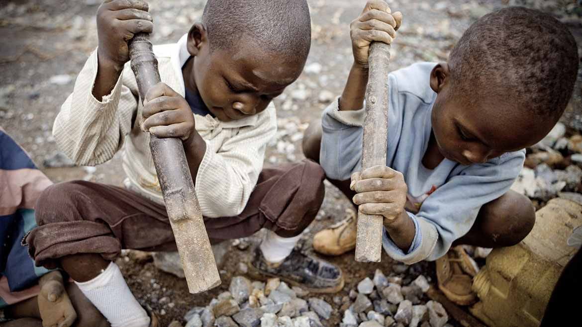 Two small Congolese boys squat in the dirt breaking cobalt ore.