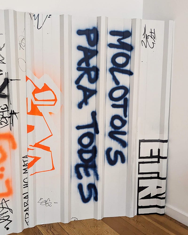 A photo of an artwork that is a spray painted white metal wall. At the center, it reads "molotovs para todes" in dark blue. A photo of an artwork that is a spray painted white metal wall. At the center, it reads "molotovs para todes" in dark blue.