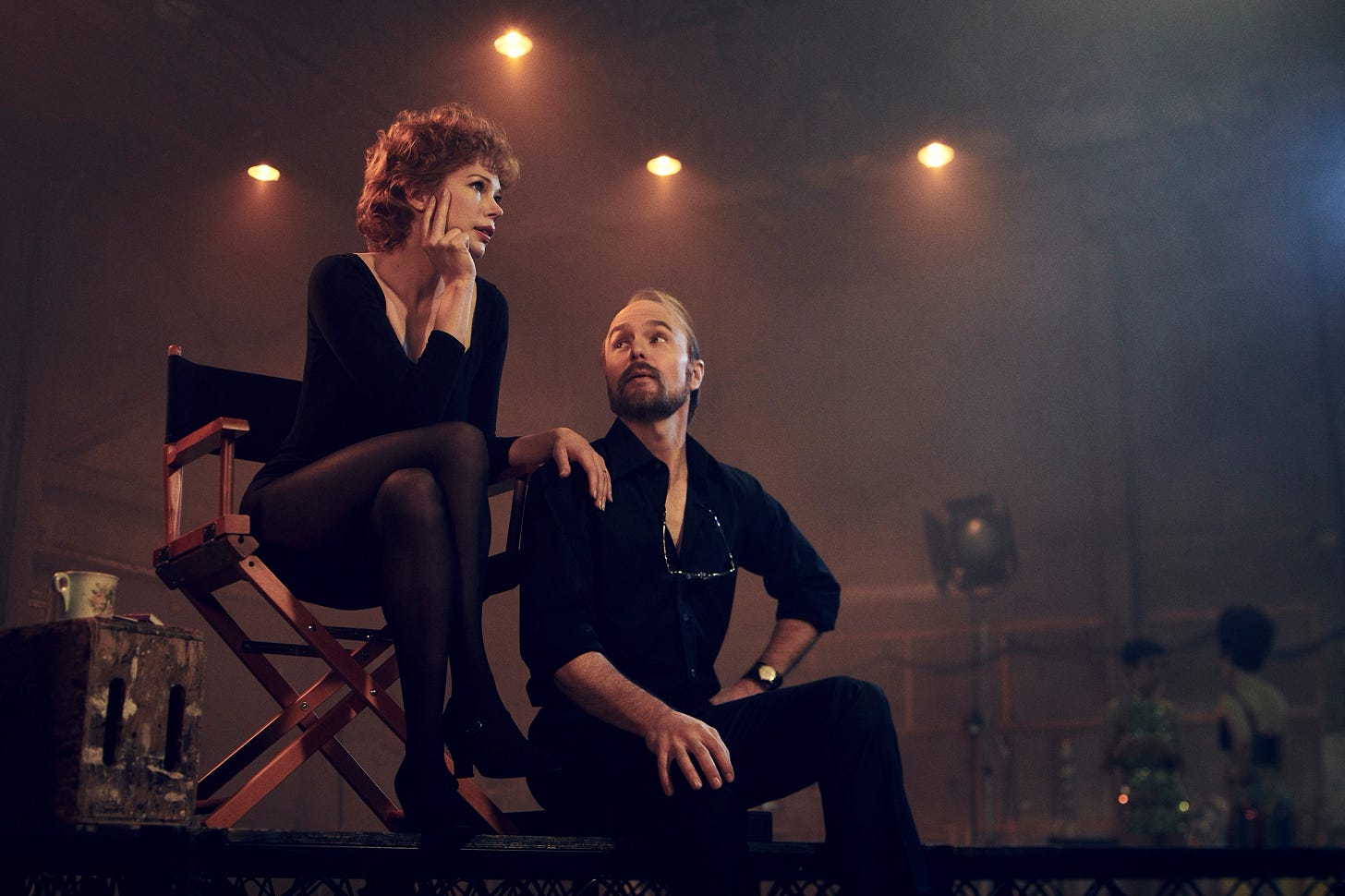 A white woman in a black leotard, played by Michelle Williams, sits in a director's chair, her hand resting on the shoulder of a white man in a black shirt and pants sitting on the floor, played by Sam Rockwell