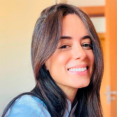 Mulher branca, de cabelos longos e negros, sorrindo; usa um jaleco branco Mulher branca, de cabelos longos e negros, sorrindo; usa um jaleco branco