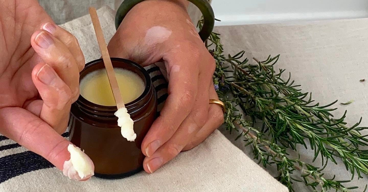 A jar or deodorant with hands holding the pot, and some product on a wooden stick and index finger. Beside this is a bunch of rosemary.