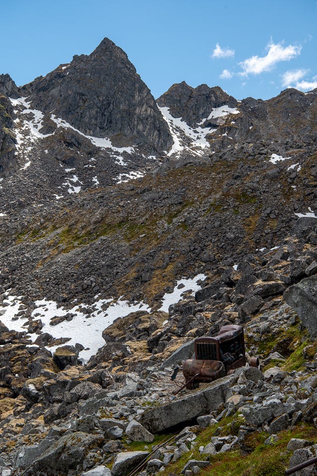 A rusted piece of old mining machinery, roughly the size of an engine block with a rounded tank on its side, sits abandoned among grey talus. Behind it, a boulder slope rises to a sharp dark peak flanked by snow-filled gullies under a blue sky. Weathered timbers and scattered iron lie among the rocks nearby. A rusted piece of old mining machinery, roughly the size of an engine block with a rounded tank on its side, sits abandoned among grey talus. Behind it, a boulder slope rises to a sharp dark peak flanked by snow-filled gullies under a blue sky. Weathered timbers and scattered iron lie among the rocks nearby.