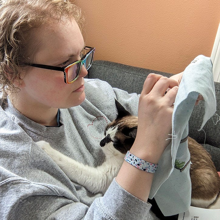 Kate from Two Little Kits trying to stitch while her cat lays on her chest happily. Kate from Two Little Kits trying to stitch while her cat lays on her chest happily.
