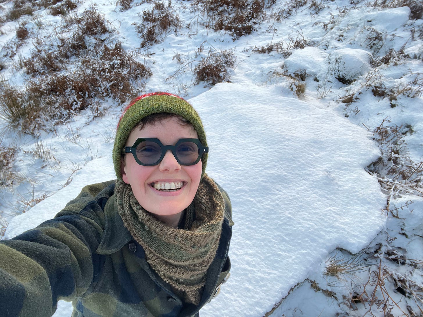 A selfie of Katie, a white person with tinted glasses grinning up at the camera, standing on a rocker covered in powdery snow. They are wearing a hand knitted hat and shawl, and checked coat and a big smile. A selfie of Katie, a white person with tinted glasses grinning up at the camera, standing on a rocker covered in powdery snow. They are wearing a hand knitted hat and shawl, and checked coat and a big smile.