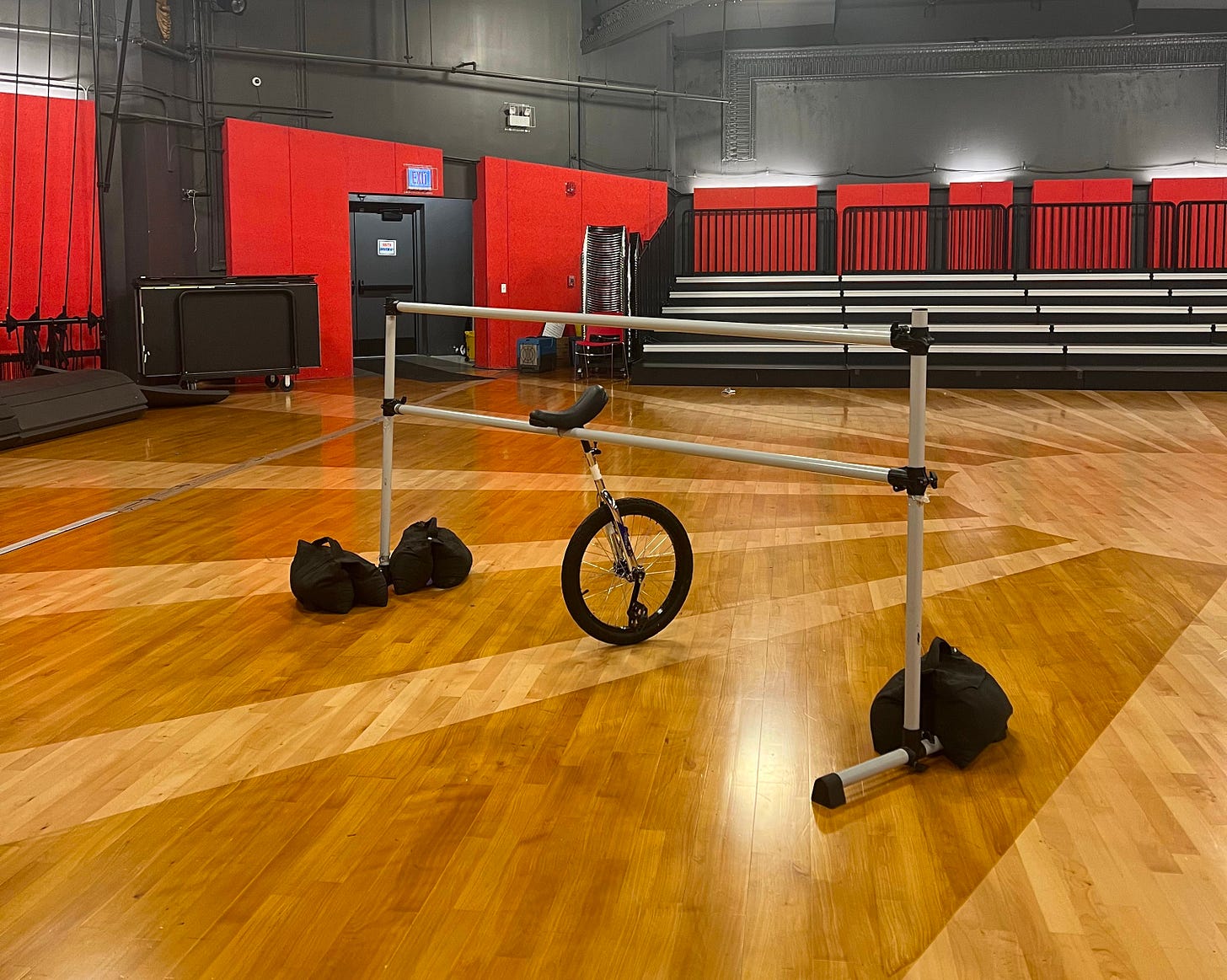 A lone unicycle in a basketball gym leaned up against a ballet-type pole with sandbags to hold it down