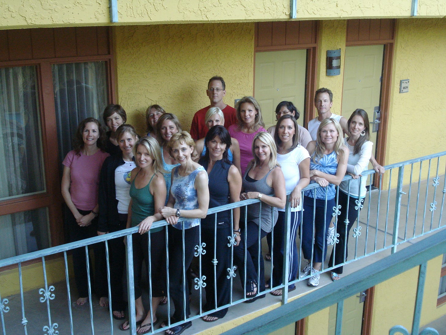 a photo of a dozen participants in a Martha Beck training including the author on a hotel balcony with terrible composition. The stuccoed walls are yellow and the banister is turquoise and adorned with flour de lis