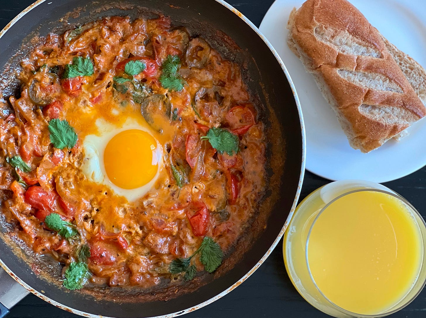 A pan of creamy shakshuka, made by Liat Portal for Foodie Disorder, featuring a golden egg yolk in a spiced tomato and pepper sauce, garnished with fresh cilantro. The dish is served in a black pan alongside a crisp French baguette on a white plate and a glass of fresh orange juice, creating a warm and inviting meal. Captured for The Liat Show, this photo embodies the comforting essence of home-cooked shakshuka.