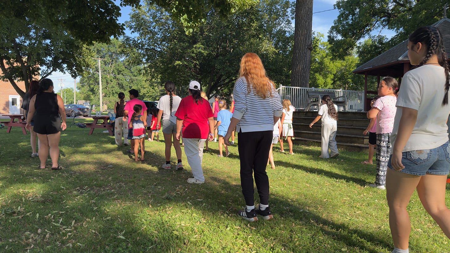 A group of kids and adults line dancing in the park.