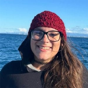 20260302_karlavieiramorato Mulher de gorro vermelho, cabelo escuro comprido e óculos. Foto tirada em frente ao mar ou a uma lagoa