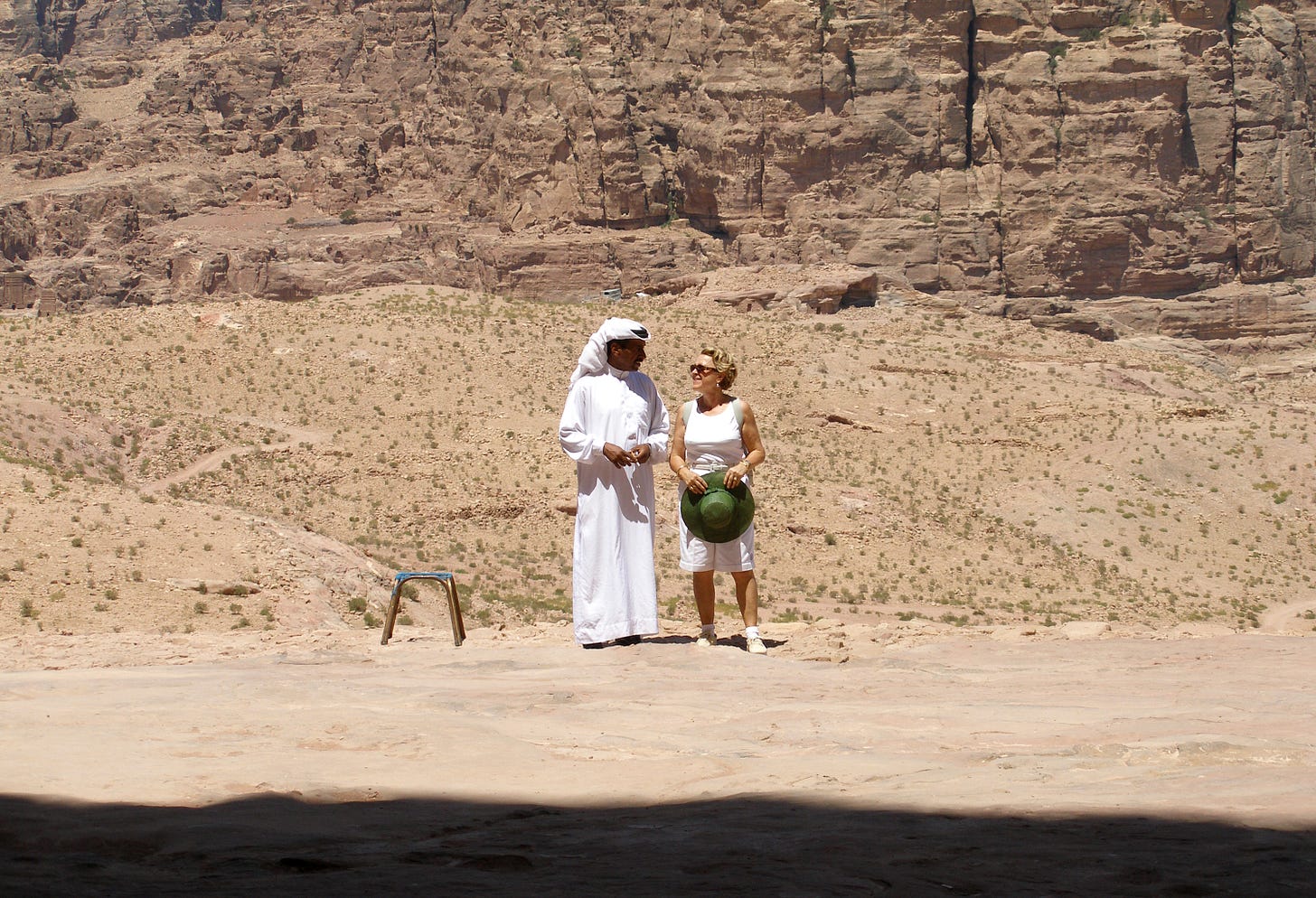 A man in a white thawb and headpiece talks to a woman in a white tank top and shorts carrying a green hat. They are surrounded by desert and cliffs.