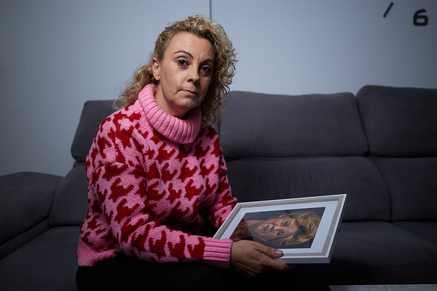 Una donna bionda con un maglione rosa e rosso siede su un divano grigio e tiene in mano una cornice bianca con la foto di una donna. Una donna bionda con un maglione rosa e rosso siede su un divano grigio e tiene in mano una cornice bianca con la foto di una donna.
