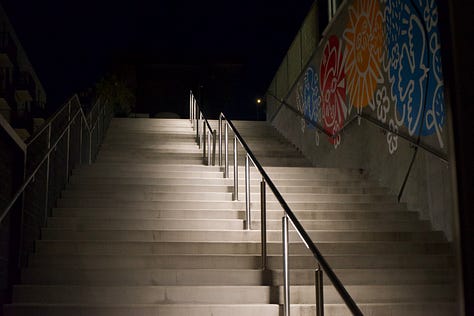 A cinematic rooftop night sequence captured in low light: orange roses glowing under warm interior lamps, reflections across glass obscuring a woman’s face as she lifts the bouquet, illuminated concrete stairs rising into shadow, a white poodle leaning from a car window through city light flares, a motion-blurred skyline beneath a crescent moon, and a circular fire pit burning at the center of a table while hands gather around it. The composition prioritizes atmosphere, reflection, and negative space over direct portraiture.