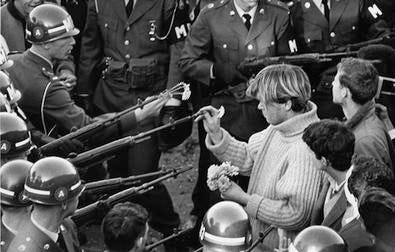 A black and white photo of a few young people on the right, surrounded by military people in uniform and on the left soldiers wearing helmets pointing guns at them. One young person in a white sweater holds up a flower and is putting into the barrel of a gun. A black and white photo of a few young people on the right, surrounded by military people in uniform and on the left soldiers wearing helmets pointing guns at them. One young person in a white sweater holds up a flower and is putting into the barrel of a gun.
