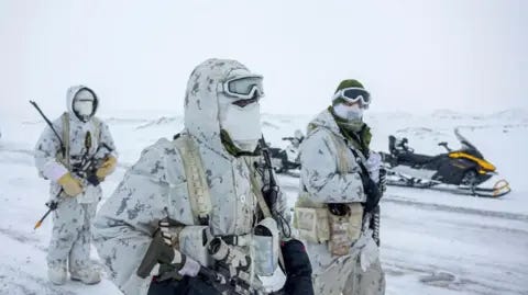 Reuters An image showing three Canadian military members in a snowy landscape, wearing white camouflage uniforms, face covers, gloves and holding various types of ammunition as they walk towards the right of the frame. Behind them is a parked snowmobile. 