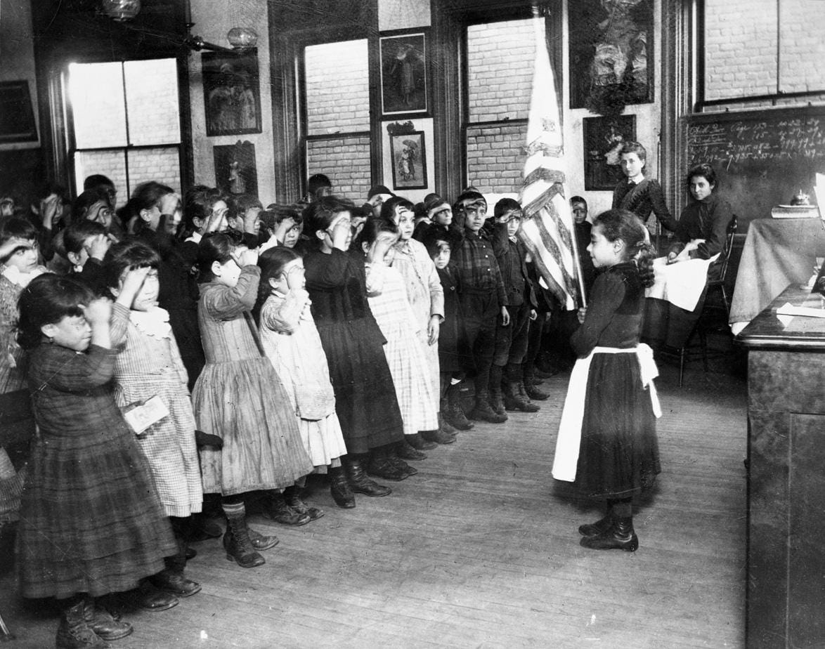 [Black and white photograph of a classroom of elementary-age schoolchildren reciting a pledge to the U.S. flag. Most students are saluting the flag with their hands raised to their brows in a military-style salute. One student stands in the front of the r