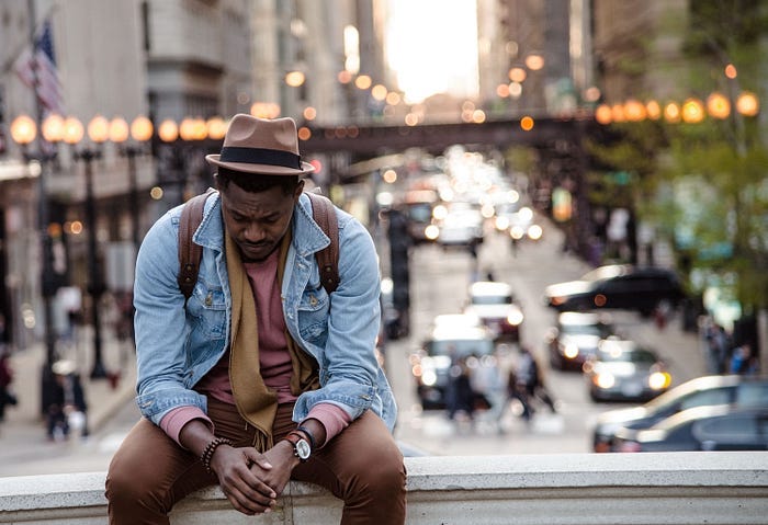african-american man sitting on wall ledge with street fair in background