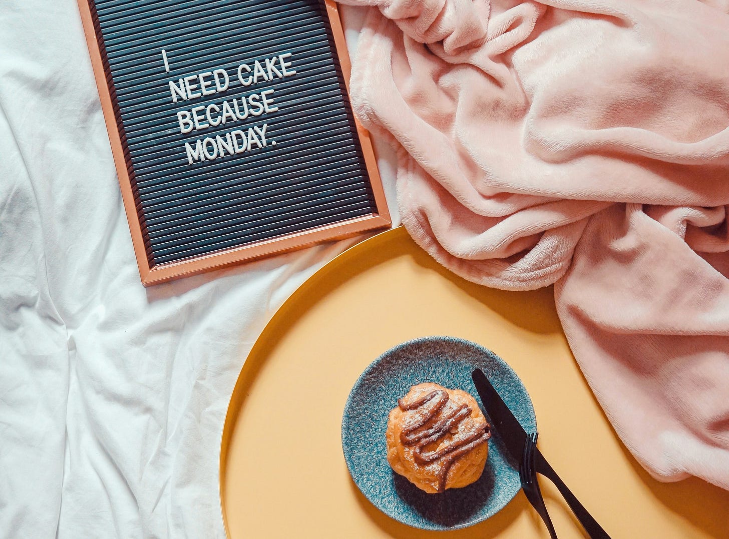 Su una coperta rosa poggia una lavagnetta su cui campeggia la scritta "I need cake because Monday" e un piattino, con una piccola torta.