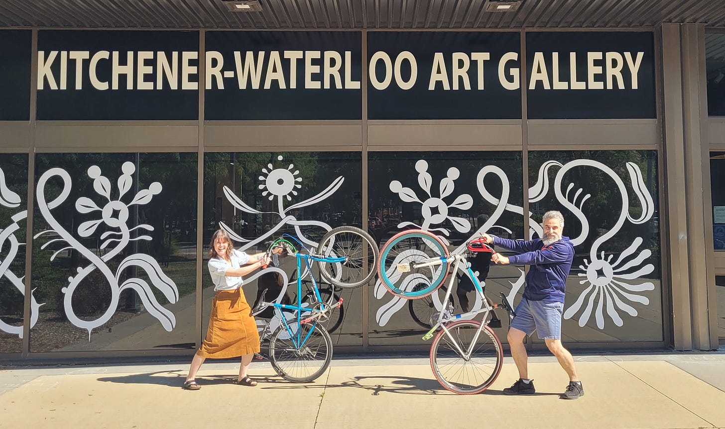 2 individuals smiling while holding their bikes upright in front of the KW art gallery