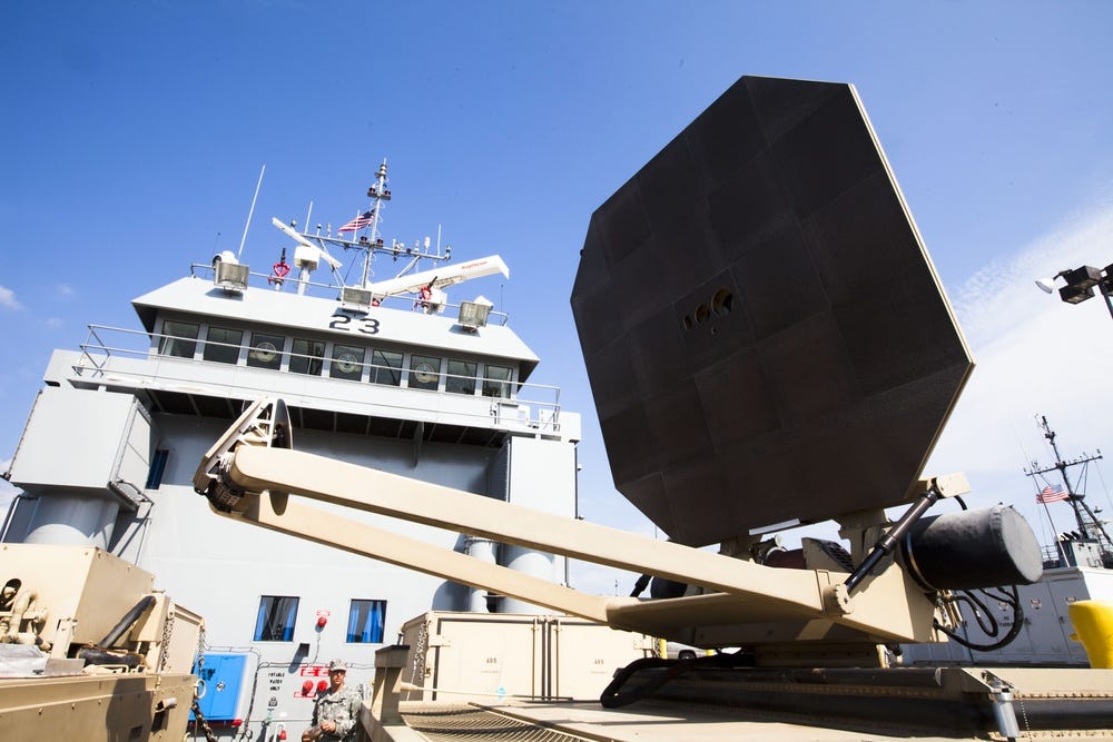 A large flat ADS antenna on a tan rotating mount sits on the deck of an Army landing craft, with the vessel's bridge behind it.