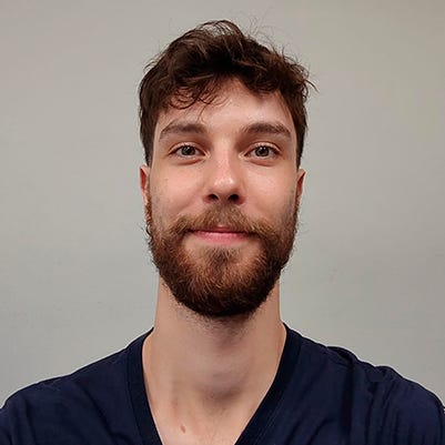 Um homem jovem com cabelo castanho ondulado e uma barba bem aparada, vestido com uma camiseta azul-marinho de gola em V, está sorrindo levemente e olhando para a câmera. Ele está posicionado no centro da imagem contra um fundo cinza claro uniforme Um homem jovem com cabelo castanho ondulado e uma barba bem aparada, vestido com uma camiseta azul-marinho de gola em V, está sorrindo levemente e olhando para a câmera. Ele está posicionado no centro da imagem contra um fundo cinza claro uniforme