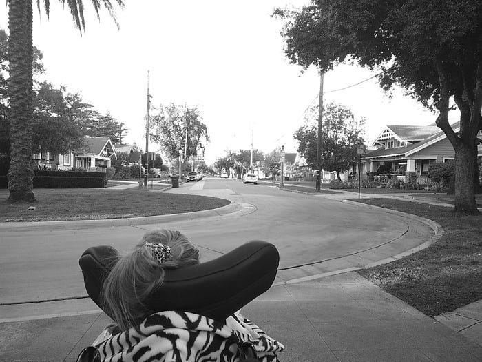 A black and white photo taken from behind a child seated in a wheelchair, wearing a zebra-print top and a hair clip, looking out at a quiet residential street lined with trees and craftsman-style homes. A black and white photo taken from behind a child seated in a wheelchair, wearing a zebra-print top and a hair clip, looking out at a quiet residential street lined with trees and craftsman-style homes.