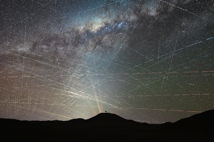 The background of this image shows a starry sky, including the bright lane of the Milky Way. Hundreds of bright streaks cross the sky in different directions, as if scratching the background natural sky behind it. The bottom of the image is occupied by the dark silhouette of a mountainous desert landscape. Atop the mountain at the centre, is a small, distant metallic dome and yellow-orange lasers shine behind it. The background of this image shows a starry sky, including the bright lane of the Milky Way. Hundreds of bright streaks cross the sky in different directions, as if scratching the background natural sky behind it. The bottom of the image is occupied by the dark silhouette of a mountainous desert landscape. Atop the mountain at the centre, is a small, distant metallic dome and yellow-orange lasers shine behind it.