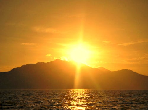 The bright yellow sun rises in a dark golden sky, behind a craggy silhouetted mountaintop, with the ocean in the foreground.