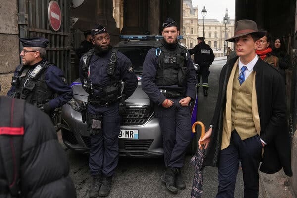 Two uniformed police officers stand beside a silver car. A person in a brown hat, black coat and vest stands with an umbrella.