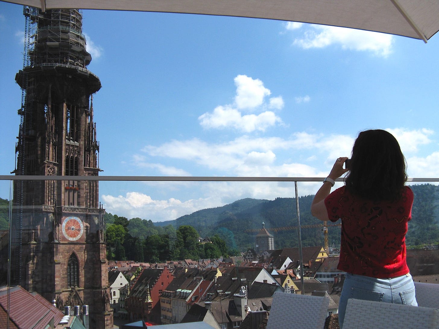 woman stand at railing of rooftop cafe taking photo of freiburg cathedral on sunny day