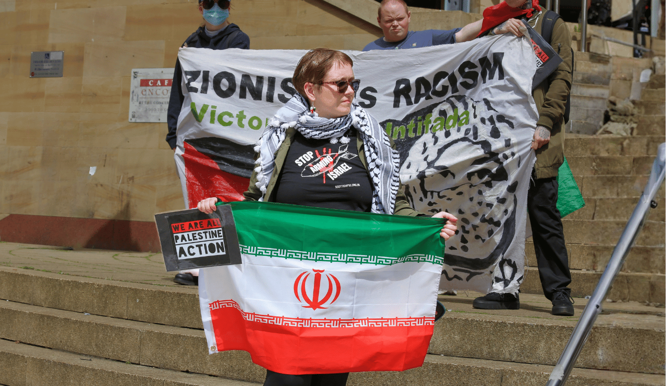 Protesters hold pro-Palestinian banners and an Iranian flag, including one reading “Zionism Is Racism—Victory to the Intifada.” Protesters hold pro-Palestinian banners and an Iranian flag, including one reading “Zionism Is Racism—Victory to the Intifada.”