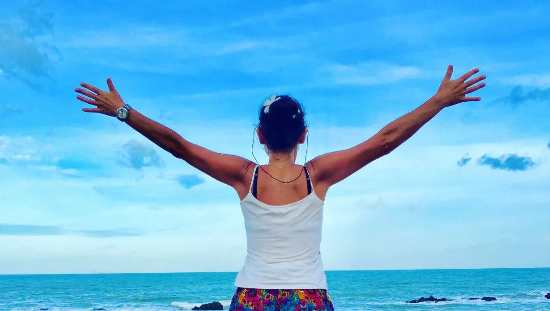A woman stands with her back to the camera, arms stretched wide open, facing a turquoise ocean under a bright blue sky in Koh Lanta, Thailand. She is wearing a white tank top and colorful floral shorts, with a flower in her hair.