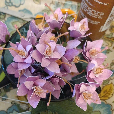Image 1 is a cup full of mauve flowers, image 2 a collection of yellow pea- style flowers and deep green spikes, Image 3 is a series of cutout green stems with thorns with some dark green leaves and pink flowers. All are made of paper and depict works in progress