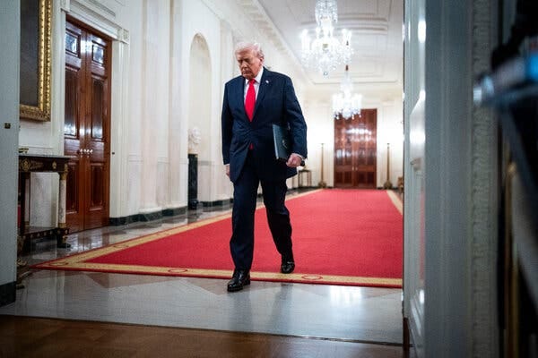 President Trump, in a suit and red tie, walking down a red-carpeted White House hallway.