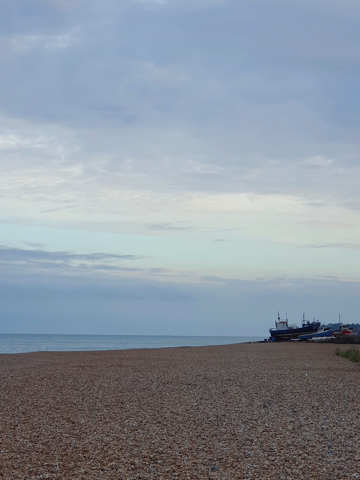 A picture of a shingle beach, some fishing boats and a vast blue sunset sky