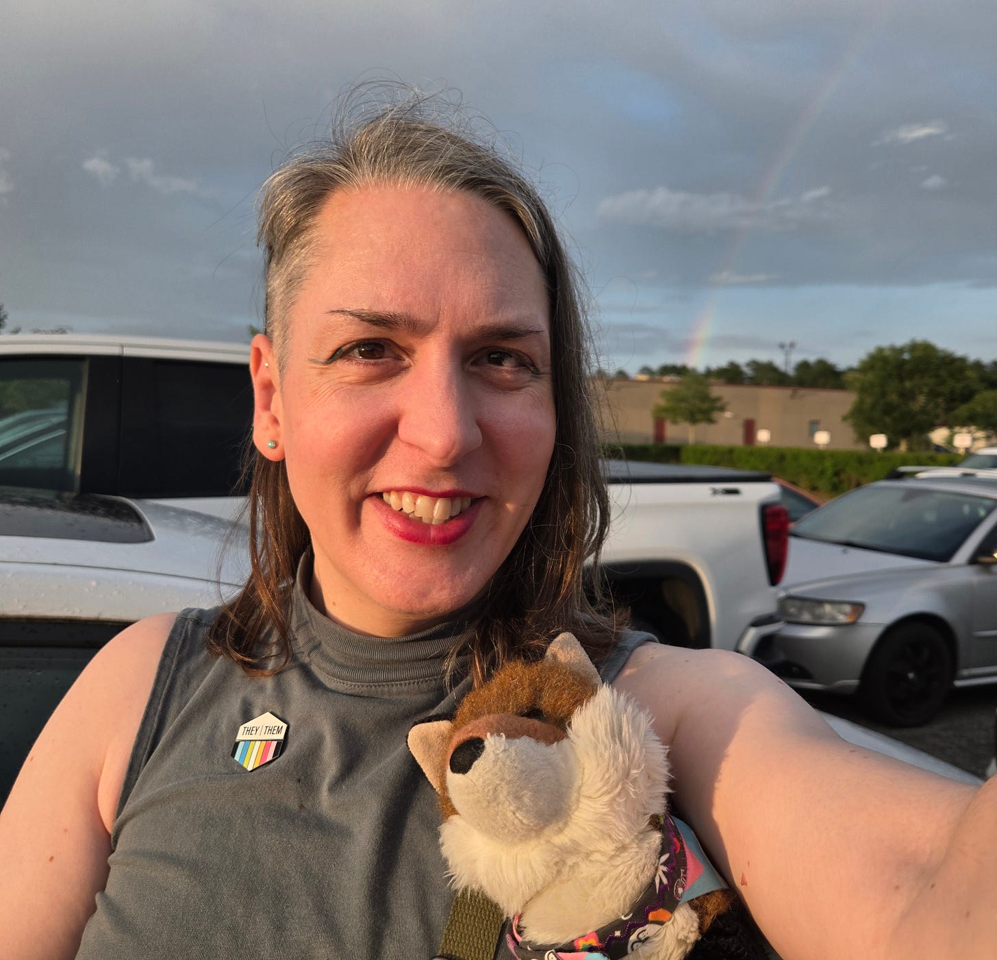 A smiling non-binary human holds a stuffed toy. They have a pin showing they/them pronouns and the queer flag colors. Behind them is a car park, and in the sky a rainbow. A smiling non-binary human holds a stuffed toy. They have a pin showing they/them pronouns and the queer flag colors. Behind them is a car park, and in the sky a rainbow.