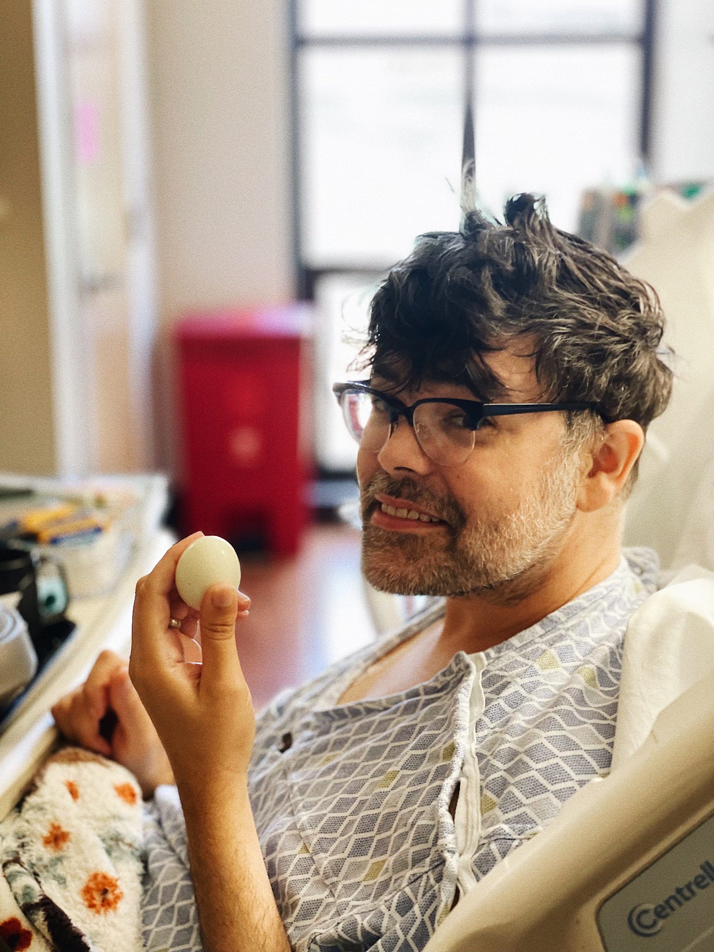 A man with tousled hair and glasses sits in a hospital bed, wearing a gown, holding a boiled egg with a playful expression.