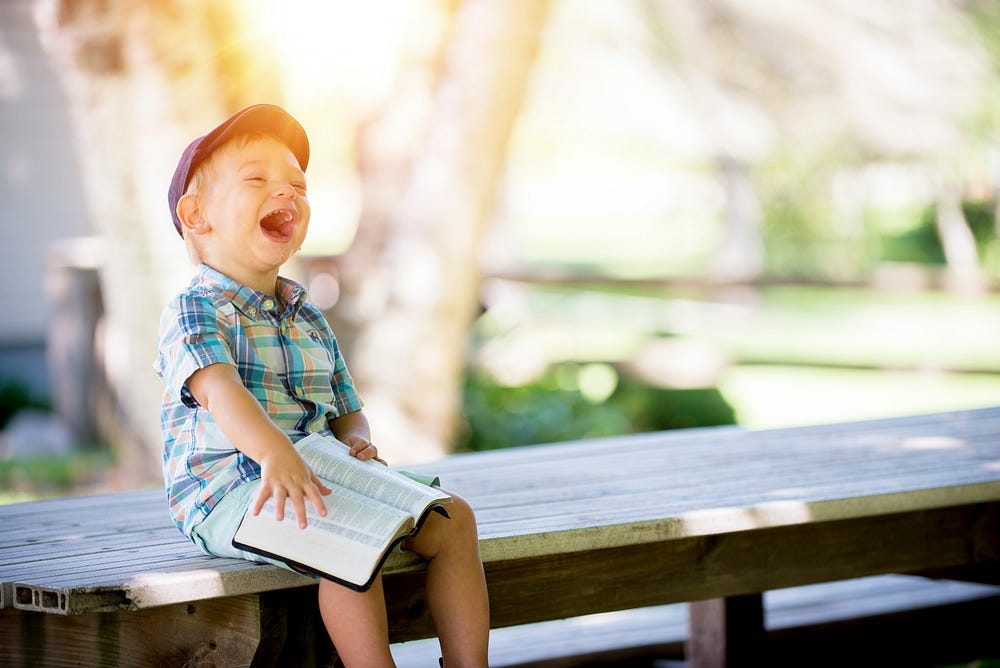 Young boy laughing while sitting on an outside bench with a Bible open on his lap.