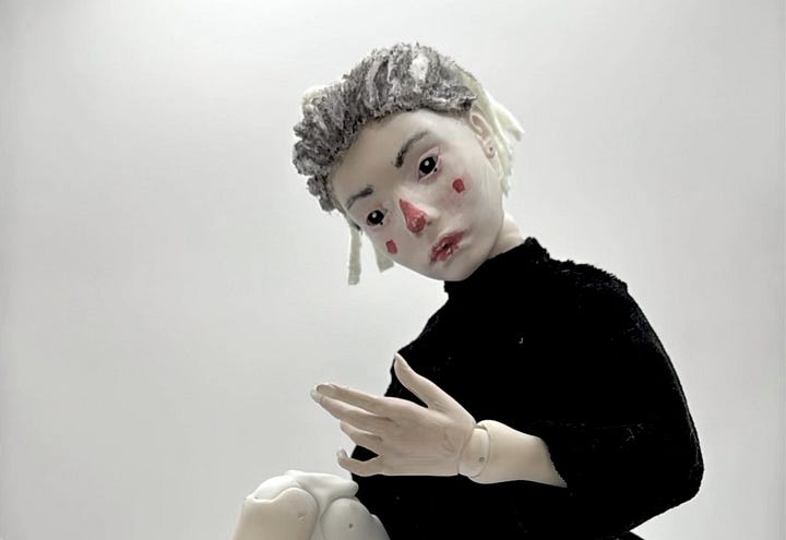 A ball jointed puppet with red dots on the cheeks and a red nose, discovering her own self