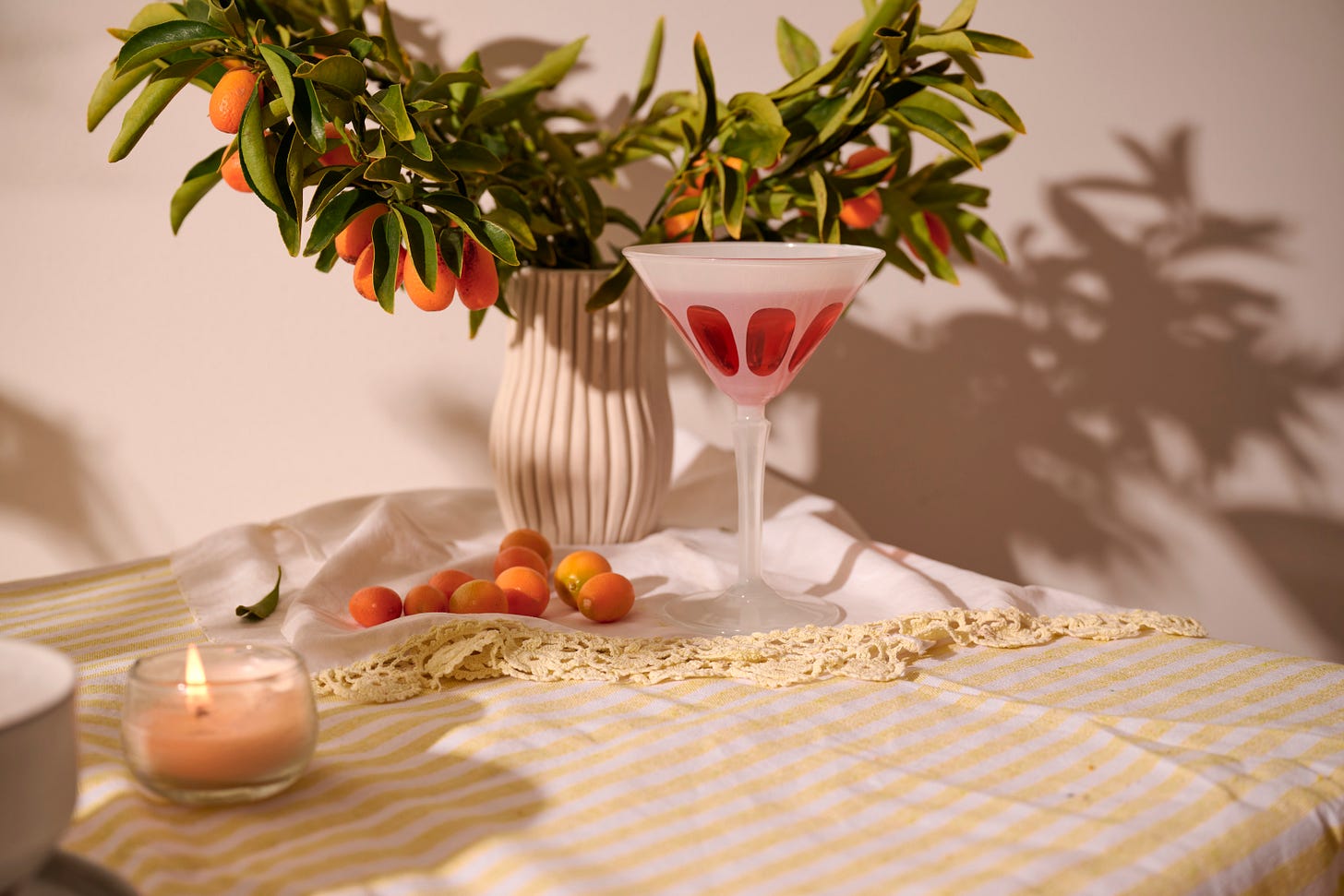 Aperol Spritz cocktail in a coupe glass with citrus garnish, styled beside fresh kumquats, a striped tablecloth, and a kumquat branch centerpiece.