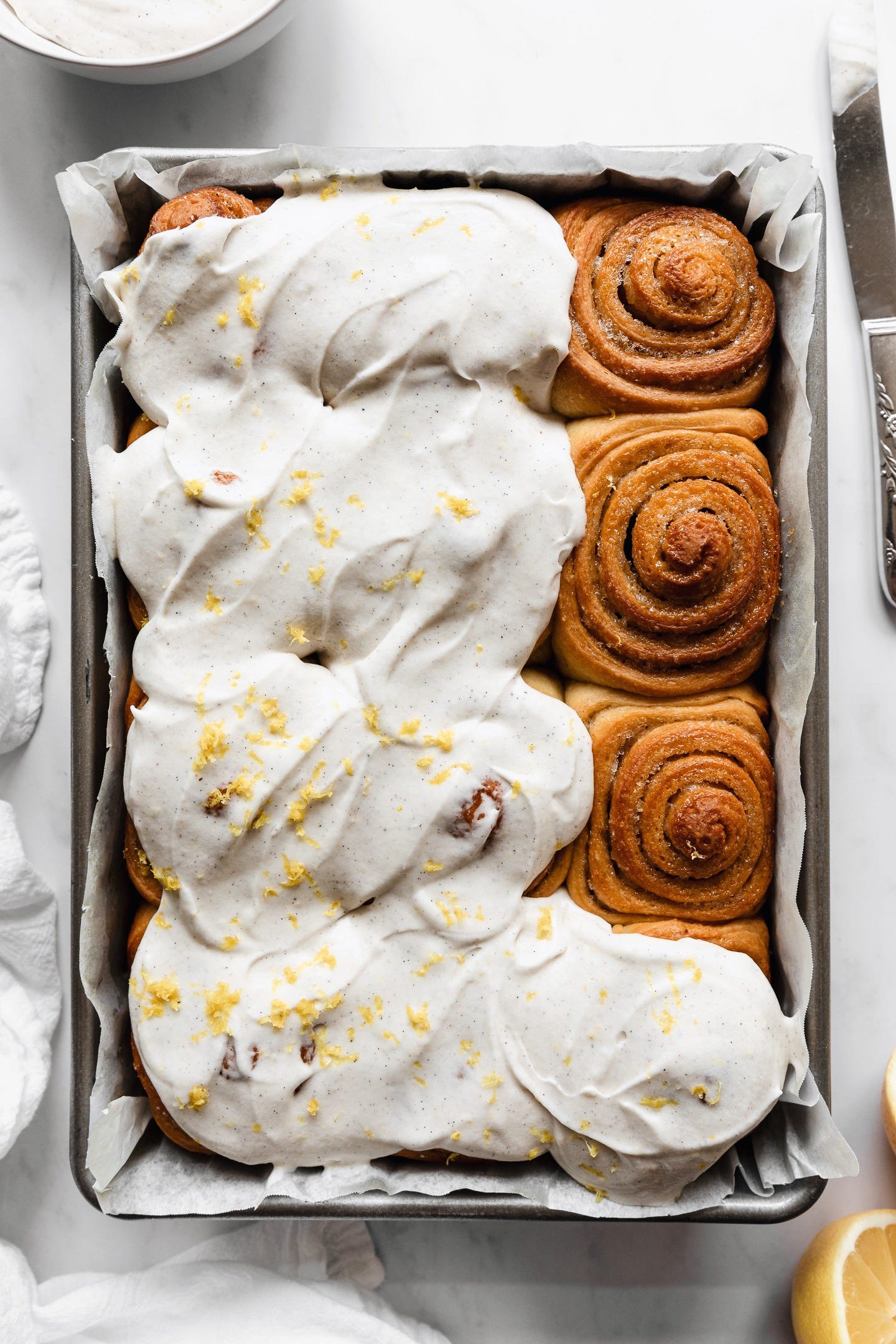 Vegan olive oil brioche buns in pan partially topped with cream cheese frosting on white counter