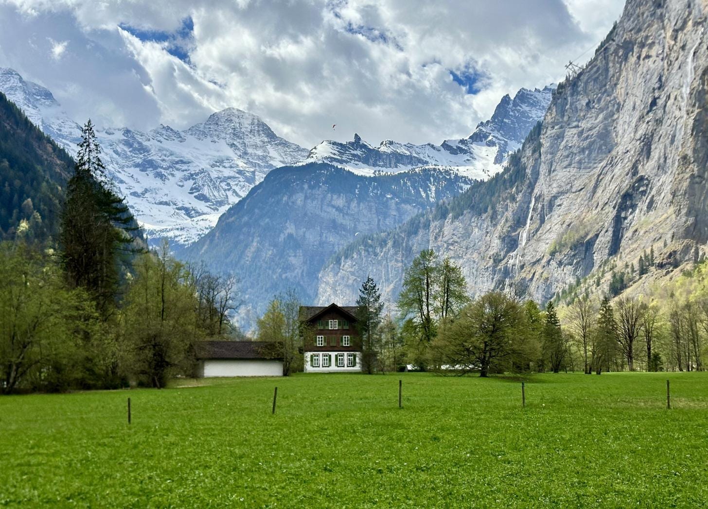 Wide view of the Lauterbrunnen valley in Switzerland with a traditional wooden Swiss chalet in a green meadow, surrounded by towering vertical limestone cliffs, snow-capped peaks, and a thin waterfall on the rock face. Wide view of the Lauterbrunnen valley in Switzerland with a traditional wooden Swiss chalet in a green meadow, surrounded by towering vertical limestone cliffs, snow-capped peaks, and a thin waterfall on the rock face.