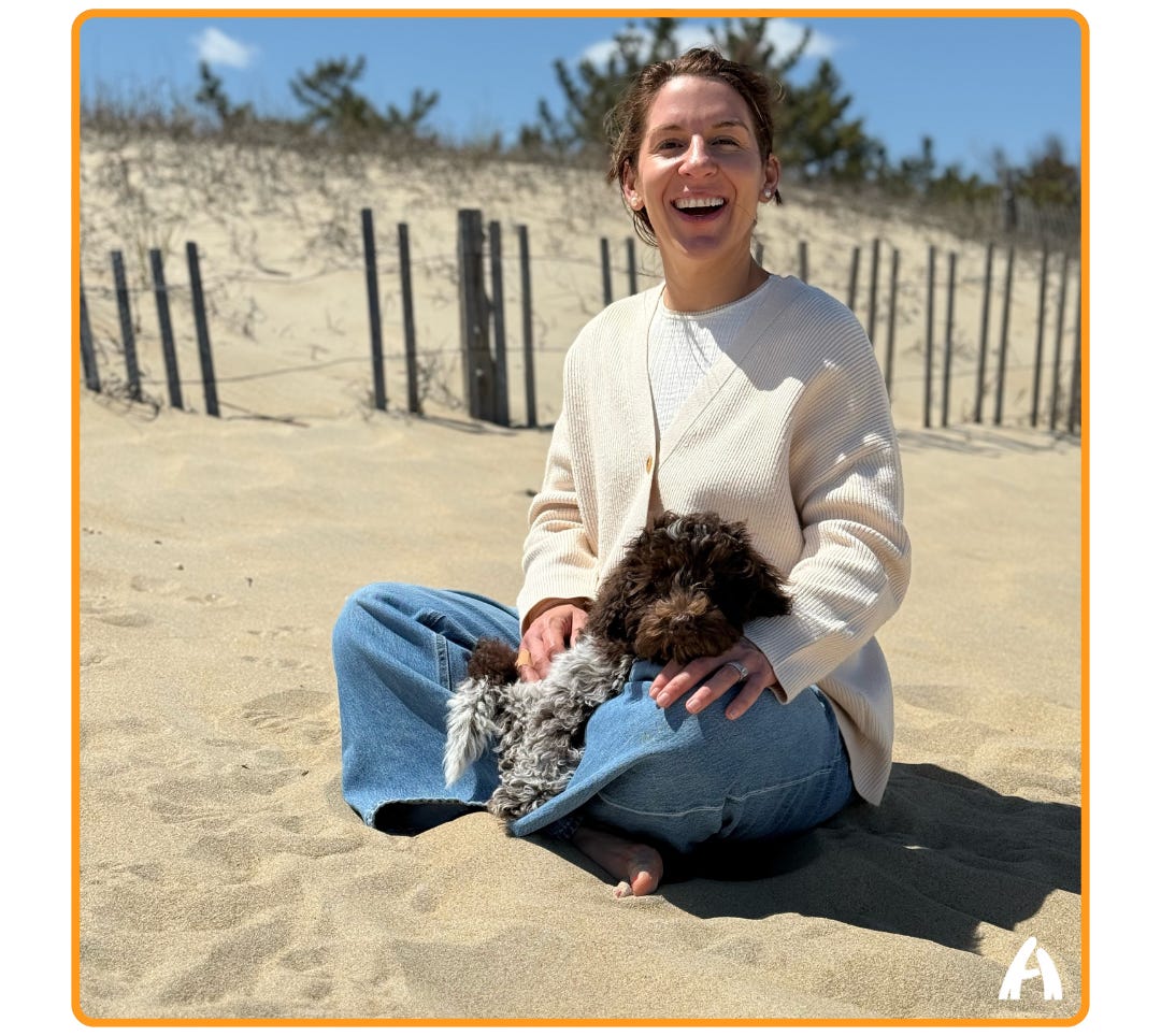 A woman sits cross-legged on a sandy beach, laughing and cradling a small curly-haired dog in her lap. She wears a cream ribbed sweater and wide-leg jeans. Sand dunes, wooden snow fencing, and pine trees are visible in the background under a blue sky.