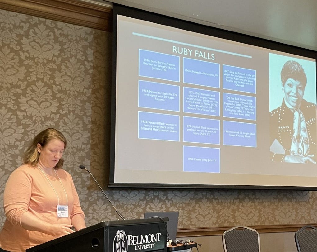 A woman at a podium presenting with a screen in the background showing text boxes and the country music star Ruby Falls.